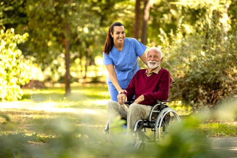 Two seniors chatting in a sunny, furnished community lounge, with staff nearby assisting another resident.